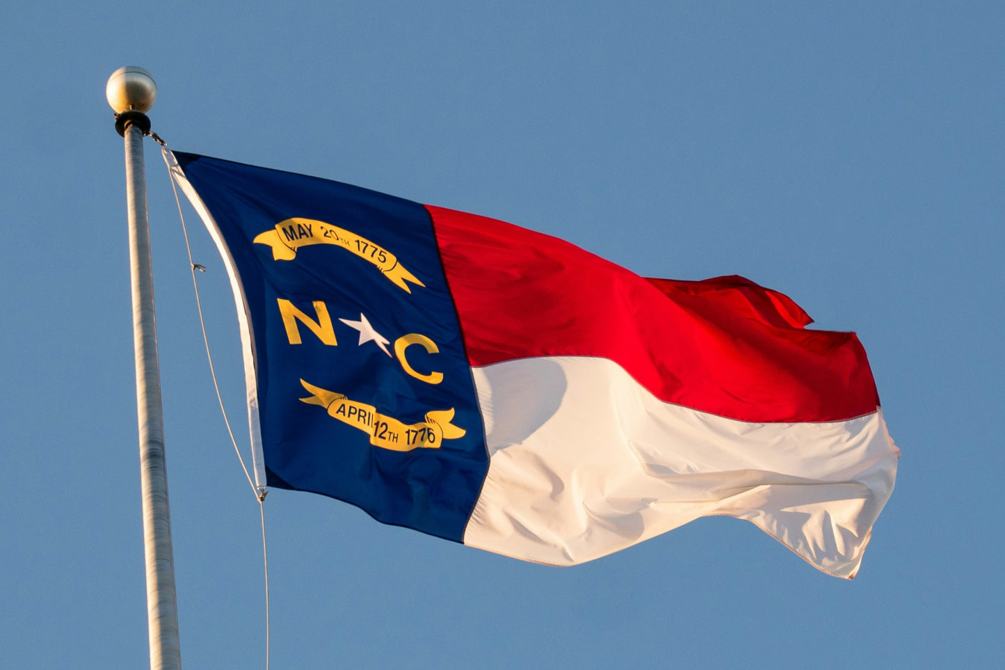 A photo of the flag of North Carolina flying on a flagpole with a blue sky in the background. 